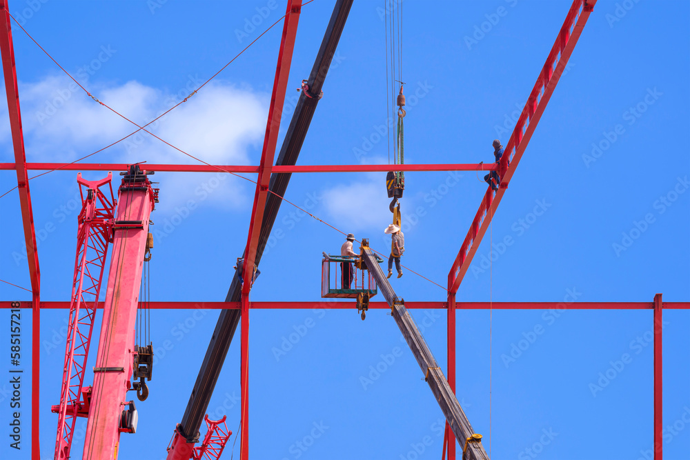 Construction workers with Crane truck are installing Sling Wire Ropes to secure metal Roof Structure of Factory Building in construction Site against blue sky background Construction workers with Crane truck are installing Sling Wire Ropes to secure metal Roof Structure of Factory Building in construction Site against blue sky background