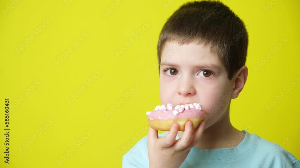 Cute 5 year old boy eats pink marshmallow donut and smiles on a yellow background Cute 5 year old boy eats pink marshmallow donut and smiles on a yellow background