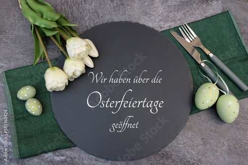 Osterdekoration mit Blumen, Ostereiern , Besteck,und dem Text  wir haben über die Osterfeiertage geöffnet auf einer Schieferplatte.
