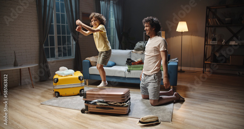 Funny asian young dad and preschooler son with curly hair are rapidly packing clothes in suitcase at home, preparing for their vacation. Family before road trip 