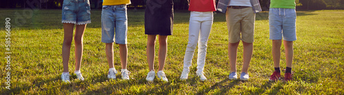 Little children in the park. Group of unrecognizable boys and girls in casual summer clothes standing in a row on green grass. Cropped shot, kids' legs. Banner or header background