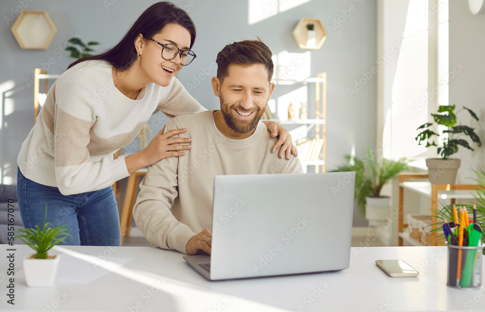 Happy young family couple using a laptop. Loving, supportive woman standing by her husband who is working on his notebook at his desk, holding him by the shoulders, looking at the computer and smiling Happy young family couple using a laptop. Loving, supportive woman standing by her husband who is working on his notebook at his desk, holding him by the shoulders, looking at the computer and smiling