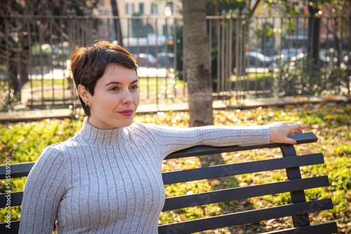 The young woman is sitting on a park bench in Rome. Beautiful woman with short hair is relaxing in the park.