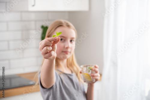 A girl drinks water with lemon and mint from a glass against the background of a bright kitchen.