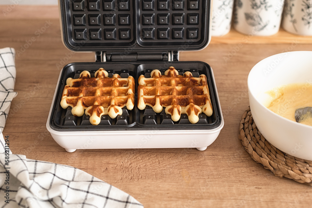 The process of making homemade Belgian waffles. A waffle iron with ready-made Belgian waffles and a bowl of homemade waffle dough on a wooden kitchen table. Cooking homemade waffles. The process of making homemade Belgian waffles. A waffle iron with ready-made Belgian waffles and a bowl of homemade waffle dough on a wooden kitchen table. Cooking homemade waffles.