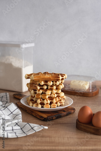 A stack of homemade Belgian waffles on a wooden kitchen table, surrounded by the ingredients to make them. Horizontal photo. Selective focus.