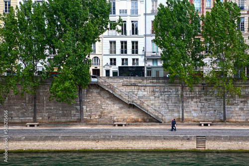 Embankment of the river Seine in Paris (France) with a cobblestone pavement, stone walls and benches, green trees, a stone staircase with iron handrails, a couple walking and a row of houses.