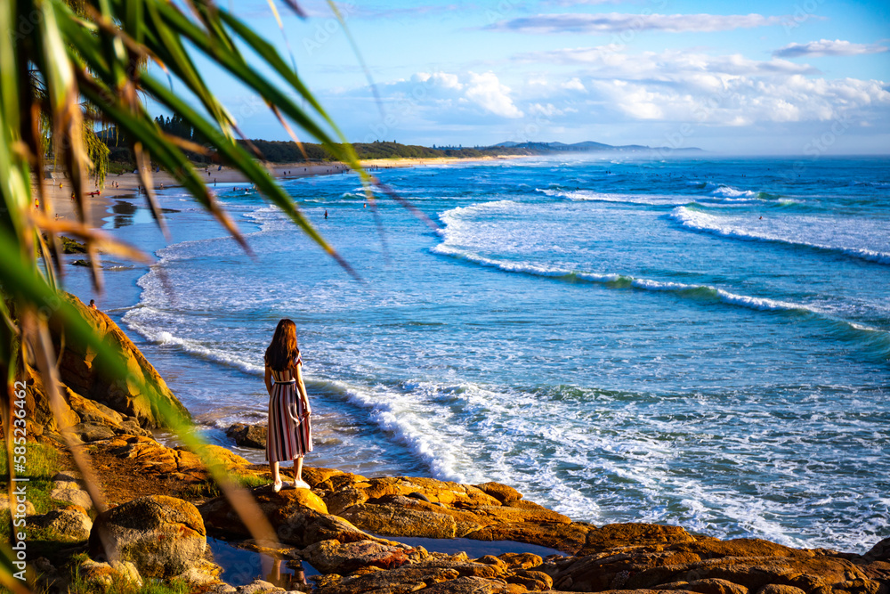 A beautiful girl in a dress stands on a rocky cliff looking at the famous Coolum beach and the Pacific Ocean and surfers catching big waves. Panorama from Sunshine Coast, Queensland, Australia