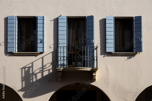 Traditional Italian building with blue wooden shutters and iron balcony creating shadows on white wall, Piove di Sacco, Padua, Italy