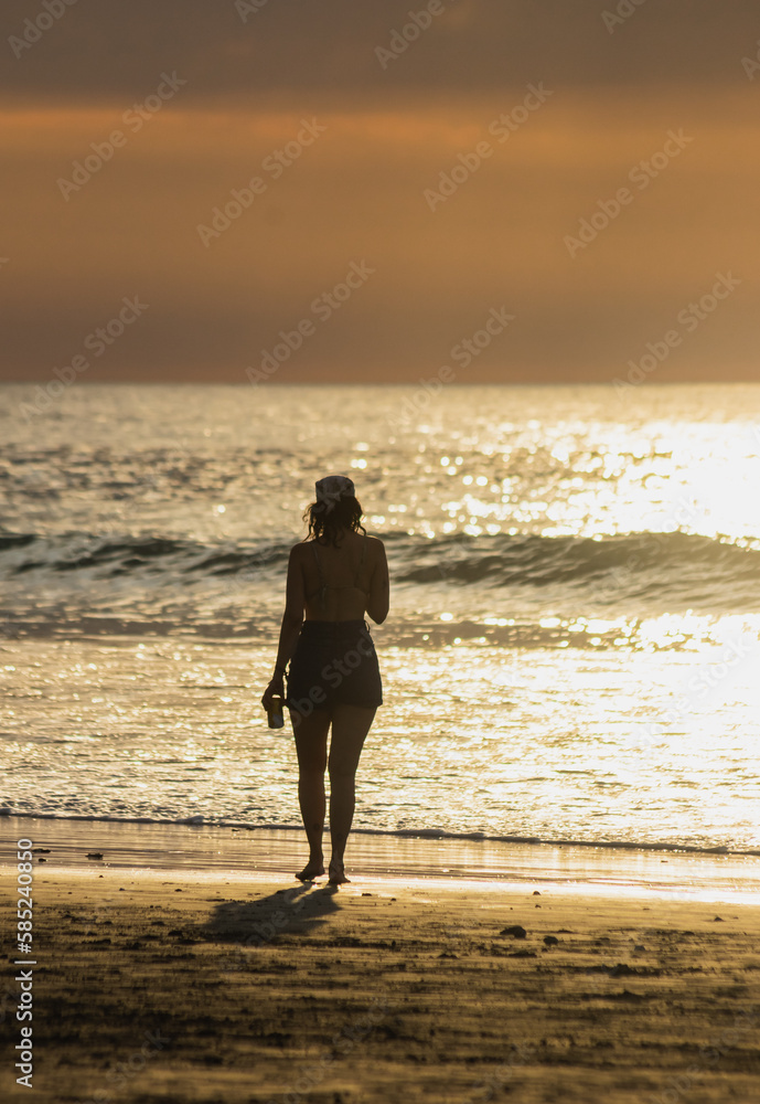 Silhouette of a girl watching the sunset on the beach near the sea and a drink in her hand with a background of the sea reflecting sunlight
