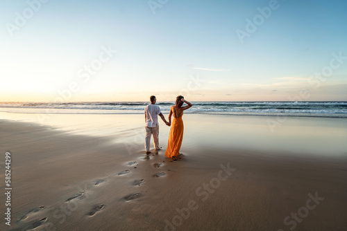 Romantic couple walking on sunset beach, enjoying evening light, relaxing on tropical summer vacation. Honeymoon. Love. Back view.