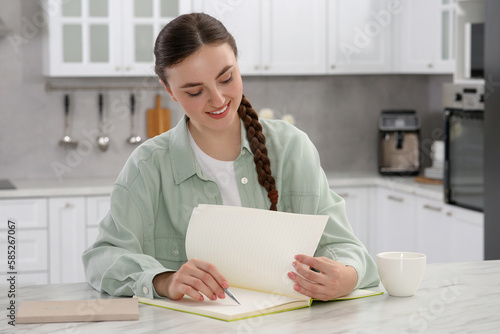 Beautiful young woman writing in notebook at white marble table indoors