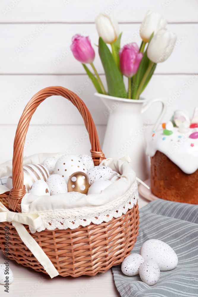 Wicker basket with festively decorated eggs, beautiful tulips and traditional Easter cake on white wooden table