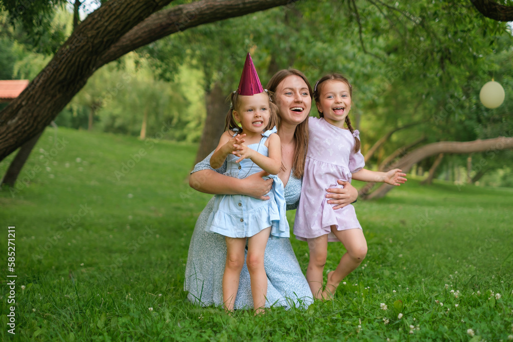 Fototapeta premium Mom hugs her daughters and laughs merrily sitting in the park on the lawn. A birthday girl with a festive hat on her head. Mothers Day.