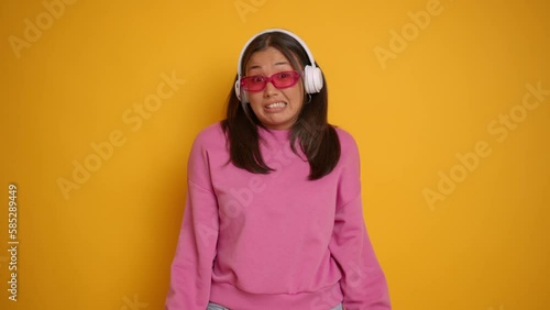 Lovely girl model wearing pink sunglasses white headphones is standing in the middle of yellow studio backdrop, smilies and shrugs shaking her head No, copy space