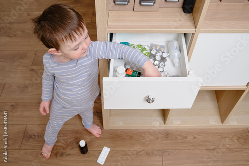 Toddler baby opened the cabinet drawer with pills and medicine. Child boy holding a pack of pills in the home living room. Kid age one year nine months