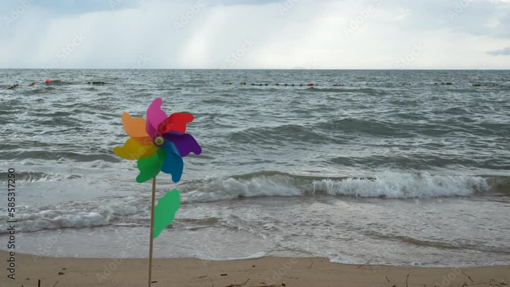 A rotated colored plastic pinwheel with a blowing wind stands on the sand by the sea against the blue smooth surface of the sea of the day. A rotated colored plastic pinwheel with a blowing wind stands on the sand by the sea against the blue smooth surface of the sea of the day.