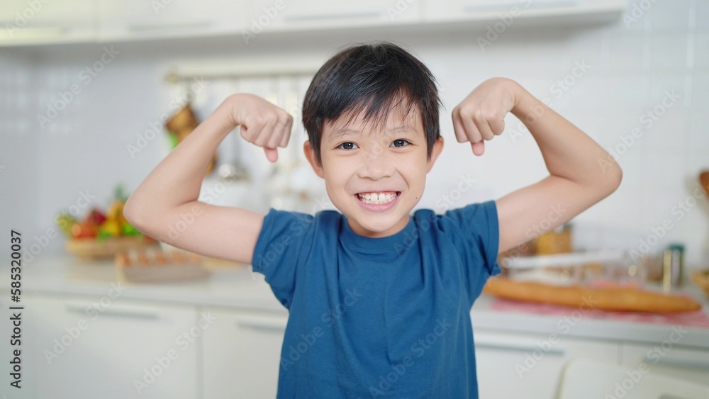 Asian little cute boy in blue shirt demonstrating his muscles or biceps at kitchen room. Boy power future and dream concept Asian little cute boy in blue shirt demonstrating his muscles or biceps at kitchen room. Boy power future and dream concept