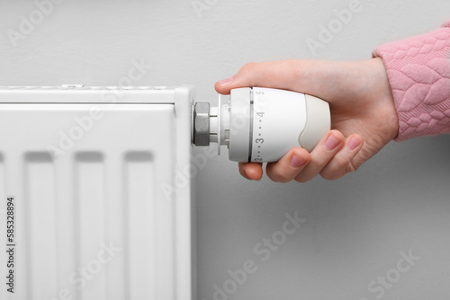 Girl adjusting heating radiator thermostat near white wall indoors, closeup