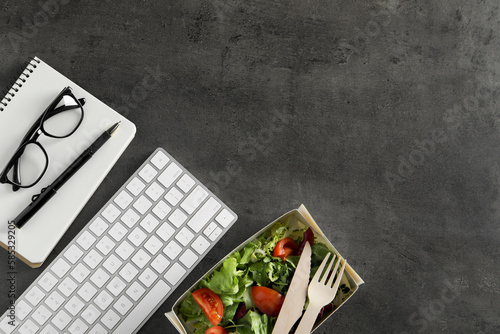Container of tasty food, keyboard, glasses, cutlery and notebook on grey table, flat lay with space for text. Business lunch