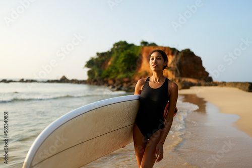 African american woman walking with surfboard on ocean beach. Black female surfer posing with surf board. Pretty multiethnic girl goes on surfing session, enjoys sun at tropical location at sunrise.