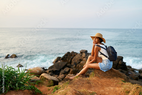 Multiethnic woman in straw hat enjoys tropical vacation sitting on rocky cliff with sea view. Black female with backpack sightseeing on scenic location. Pretty lady tanning on island on ocean sunrise.