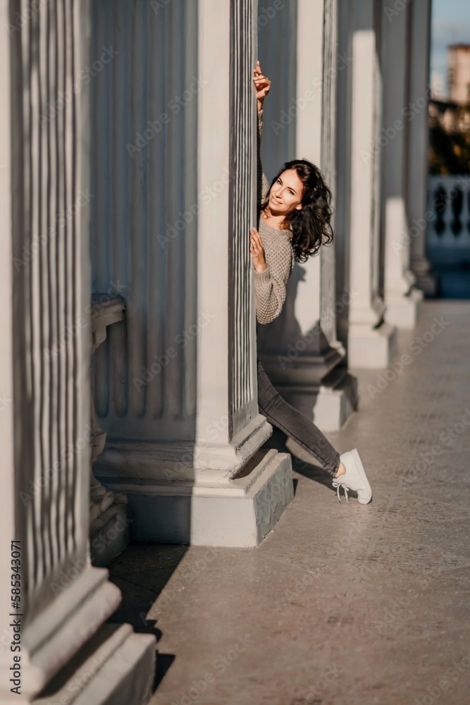 Woman building columns. An athletic woman in her 40s, dressed in a beige sweater and black jeans, poses near the pillars of a building. Walking around the city, tourism.