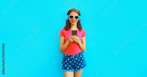 Portrait of happy smiling young woman in headphones listening to music with smartphone on blue background