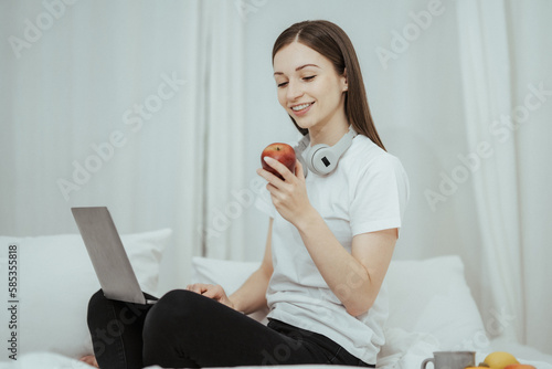 Happy woman holding apple and listening to music sitting on sofa in living room at home © Kritdanai