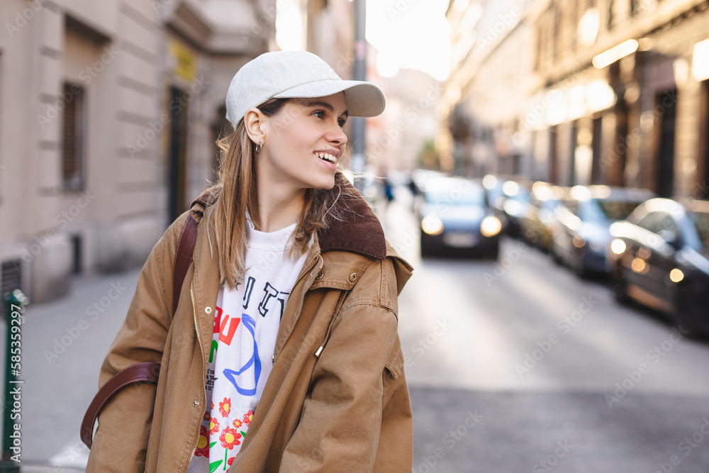 Happy girl with blonde hair in light cap and brown trench coat with handbag smiling outdoors, turn around and smiling. Stylish girl in fashion outfit walking over the city, look happy, look at side
