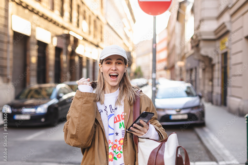 Fototapeta premium Delighted blonde girl making winner gesture and using mobile phone walking on the street. Portrait of surprised hipster girl surfing internet on mobile phone outdoors.