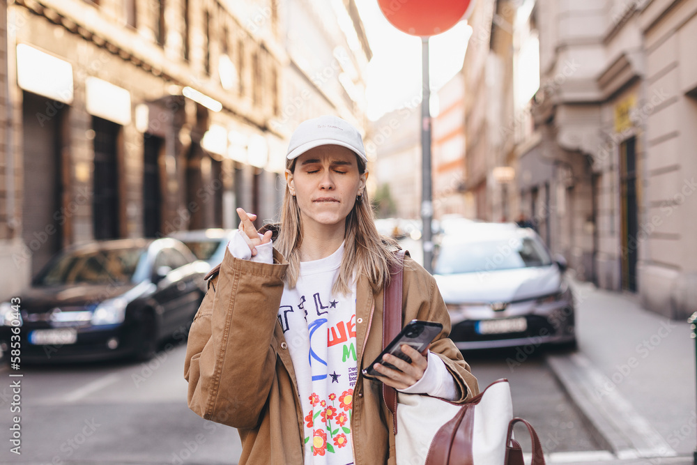 Fototapeta premium Portrait of wishful young woman in casual clothes with blonde hair, crossing her fingers, feeling nervous hold mobile phone, walking on the street. Body language.