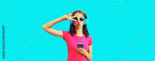 Portrait of stylish young woman in headphones listening to music with smartphone and blowing her lips sending air kiss on blue background