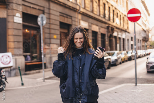 Happy smiling woman hold smartphone with excited face expression, closed eyes, unable to believe that she bet played, win in lottery, read good news, can't believe in her fortune walking outdoors.