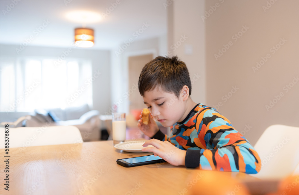 Healthy Kid having breakfast, Happy Child Boy using mobile phone watching, reading or playing games a while eating food, School Kid croissant and milk before go to school in the morning Healthy Kid having breakfast, Happy Child Boy using mobile phone watching, reading or playing games a while eating food, School Kid croissant and milk before go to school in the morning