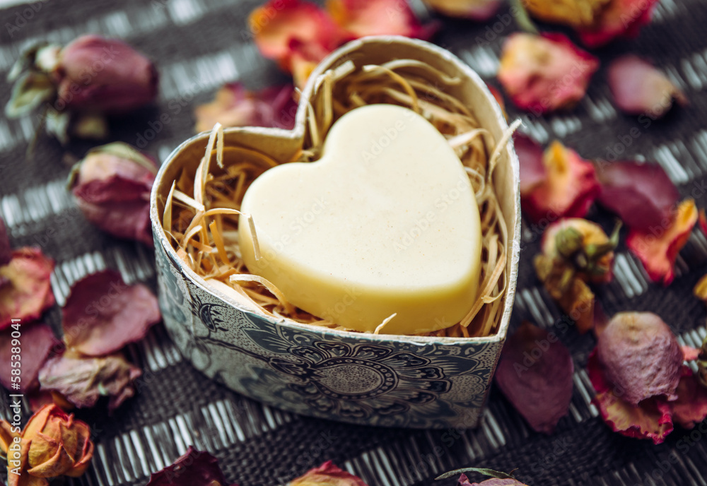 Solid Body cream bar in a heart shape gift box on black background, decorated with dry rose flower petals indoors.