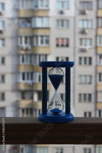 Blue Hourglass With Sand Falling Down On A Wooden Railing Of The Balcony Of A High-Rise Building 