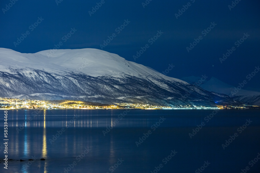 Long exposure cold night photograph of the Norwegian city of Tromsø, Northern Norway. 30 seconds. Mountains covered in snow and motion blurred water reflections of the lights.