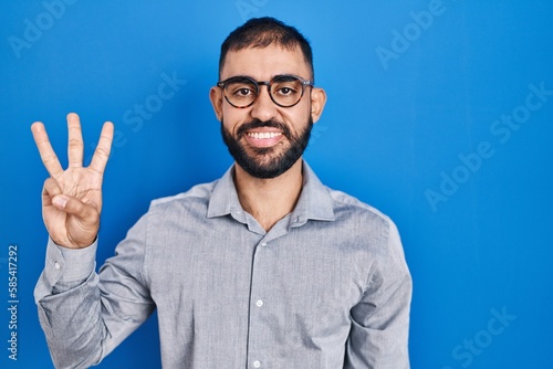 Middle east man with beard standing over blue background showing and pointing up with fingers number three while smiling confident and happy.