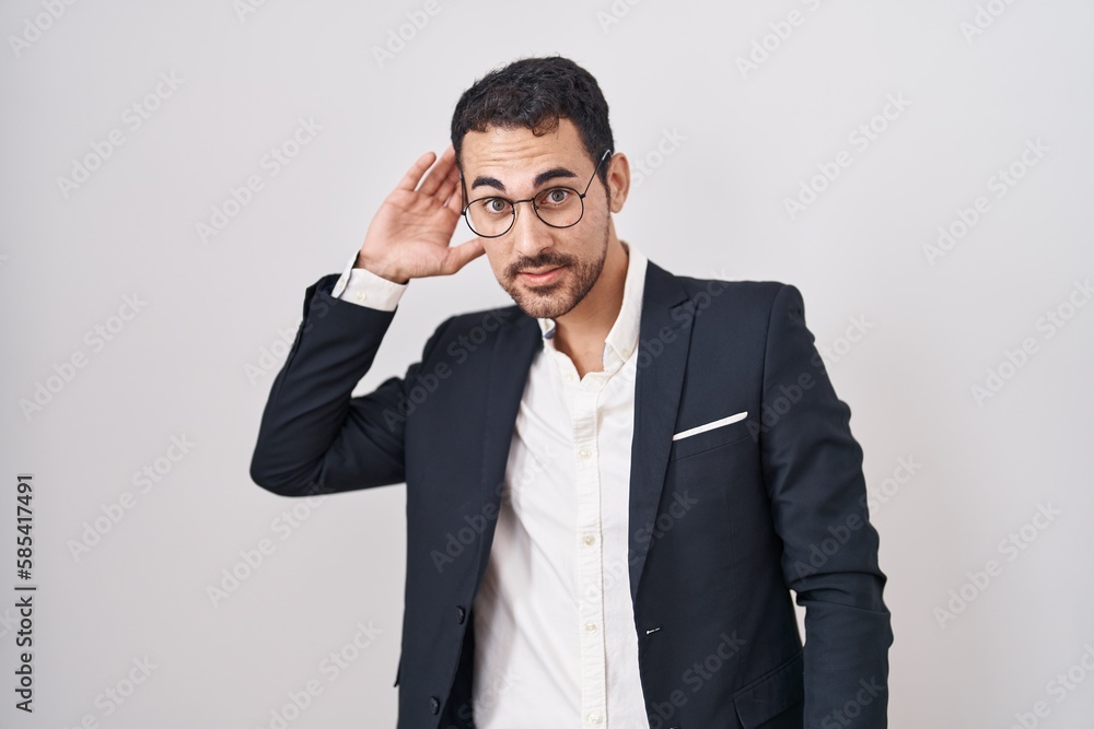Handsome business hispanic man standing over white background smiling with hand over ear listening an hearing to rumor or gossip. deafness concept. Handsome business hispanic man standing over white background smiling with hand over ear listening an hearing to rumor or gossip. deafness concept.
