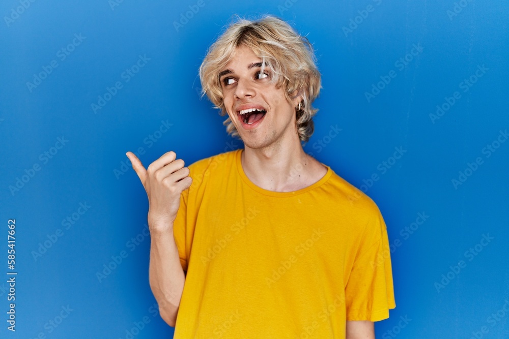 Young modern man standing over blue background smiling with happy face looking and pointing to the side with thumb up. Young modern man standing over blue background smiling with happy face looking and pointing to the side with thumb up.