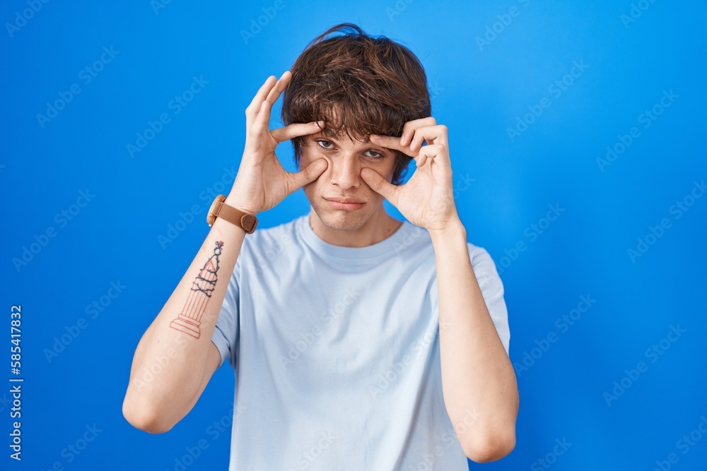 Hispanic young man standing over blue background trying to open eyes with fingers, sleepy and tired for morning fatigue Hispanic young man standing over blue background trying to open eyes with fingers, sleepy and tired for morning fatigue
