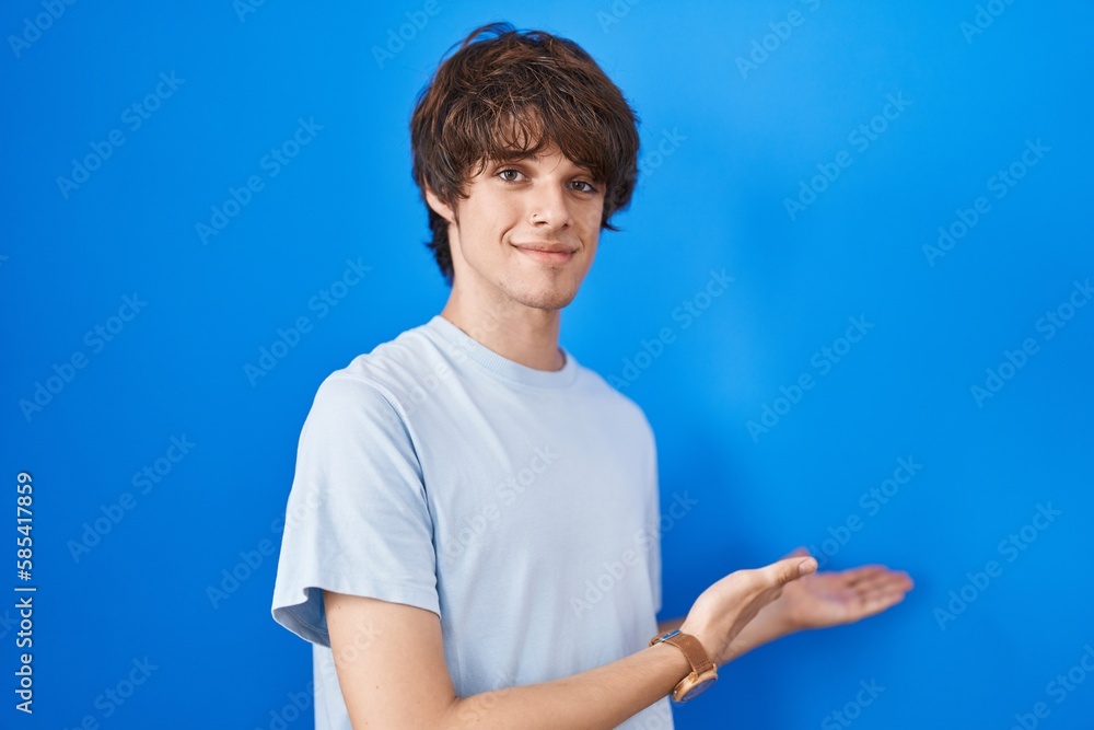 Hispanic young man standing over blue background inviting to enter smiling natural with open hand Hispanic young man standing over blue background inviting to enter smiling natural with open hand