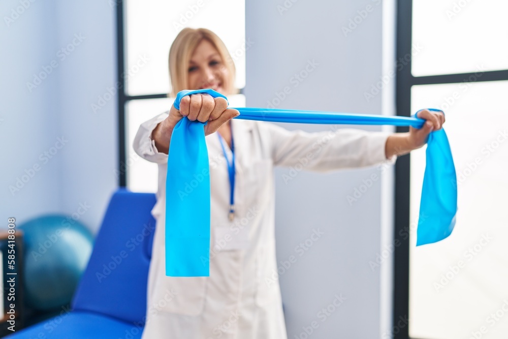 Middle age blonde woman wearing physiotherapist uniform using elastic band at physiotherapy clinic