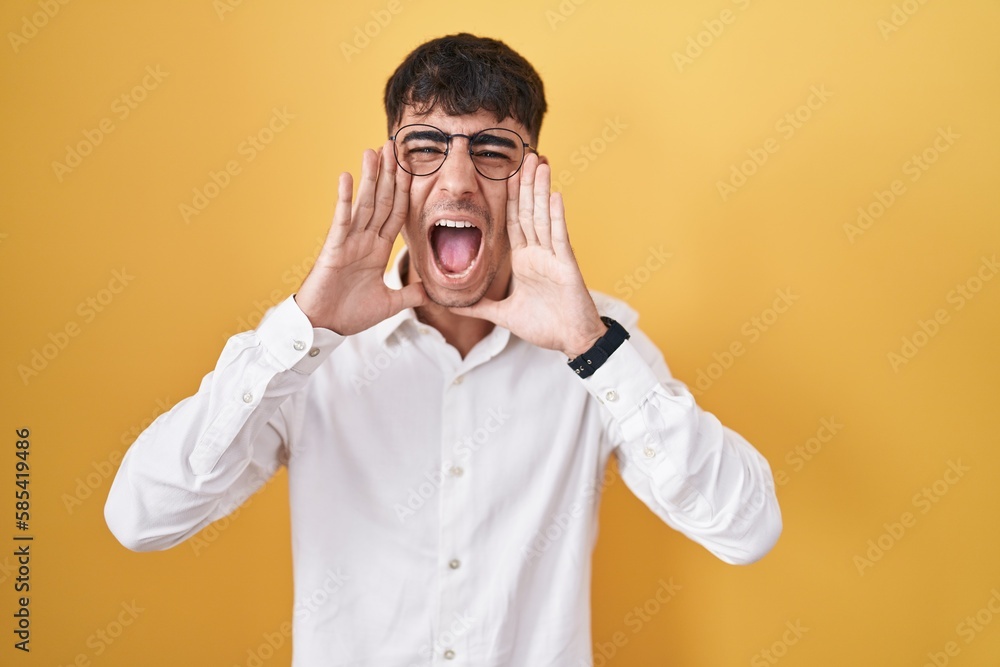 Young hispanic man standing over yellow background shouting angry out loud with hands over mouth Young hispanic man standing over yellow background shouting angry out loud with hands over mouth