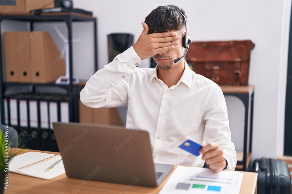 Obraz premium Young hispanic man working using computer laptop holding credit card covering eyes with hand, looking serious and sad. sightless, hiding and rejection concept