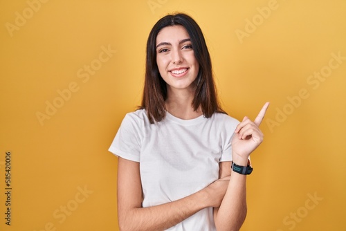 Young beautiful woman standing over yellow background with a big smile on face  pointing with hand finger to the side looking at the camera.
