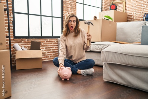 Young woman holding piggy bank and hammer moving to a new home in shock face, looking skeptical and sarcastic, surprised with open mouth