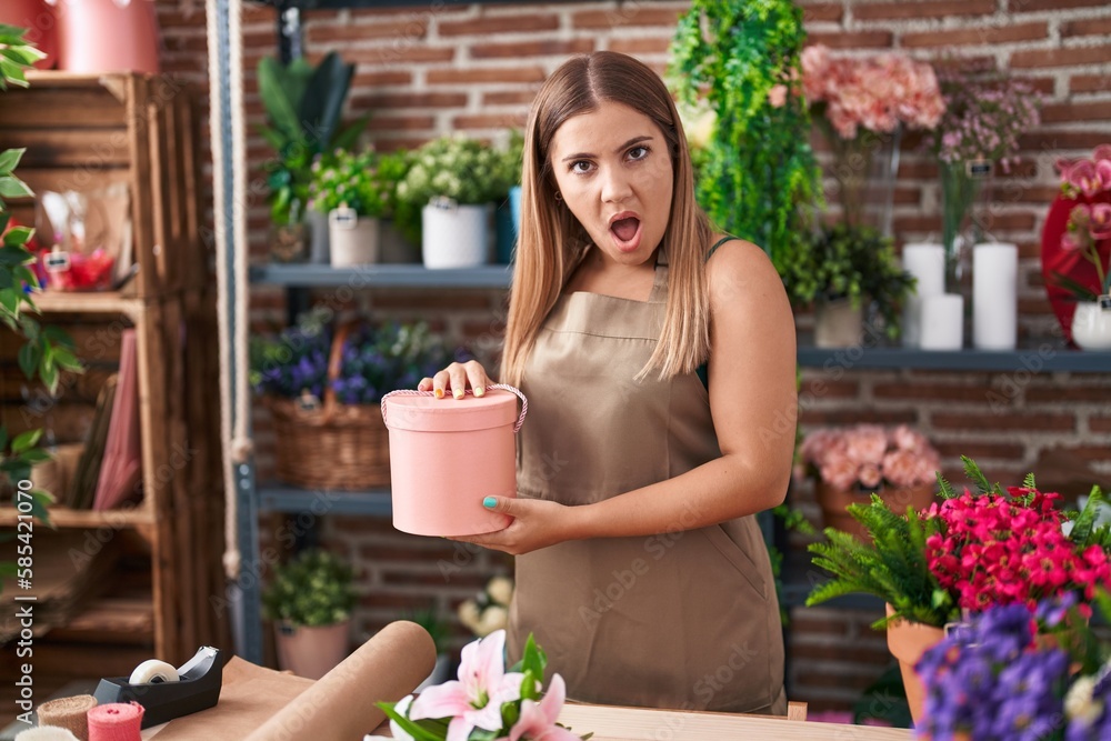 Young blonde woman working at florist shop in shock face, looking skeptical and sarcastic, surprised with open mouth