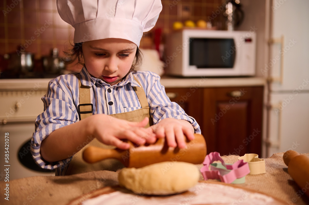 Adorable Caucasian 5-6 years child girl, little chef confectioner in cook uniform, standing at kitchen island and using rollin pin, rolls out dough on floured wooden board, prepares Easter gingerbread Adorable Caucasian 5-6 years child girl, little chef confectioner in cook uniform, standing at kitchen island and using rollin pin, rolls out dough on floured wooden board, prepares Easter gingerbread